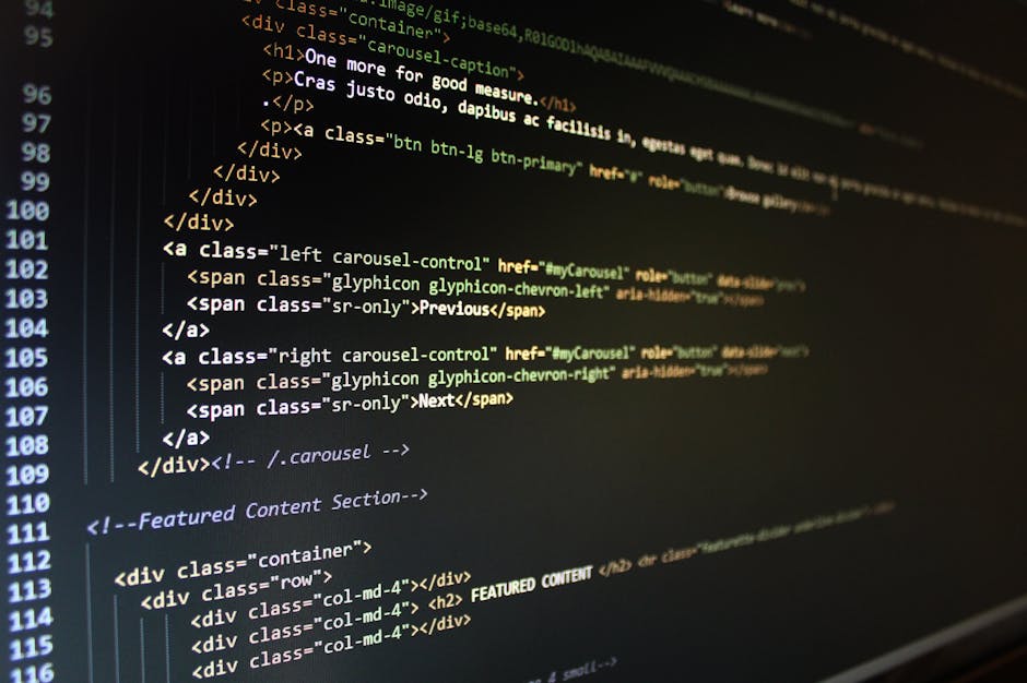 A close-up view of a person's hand using a laptop; the screen displays lines of code with syntax highlighting, including elements like tags and programming symbols. The laptop has a silver exterior wi