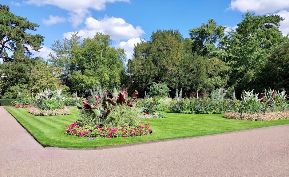 The image depicts a well-maintained garden area within Lamorbey Park, featuring lush, vibrant green grass bordered by neatly trimmed edges. In the foreground, there are flower beds filled with colourf